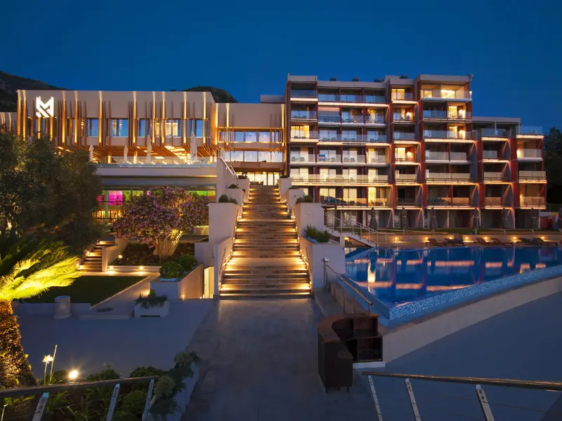 Eksterijer Maestral Resort & Casino noću sa osvetljenim stepeništem, bazenom i M logom na krovu
