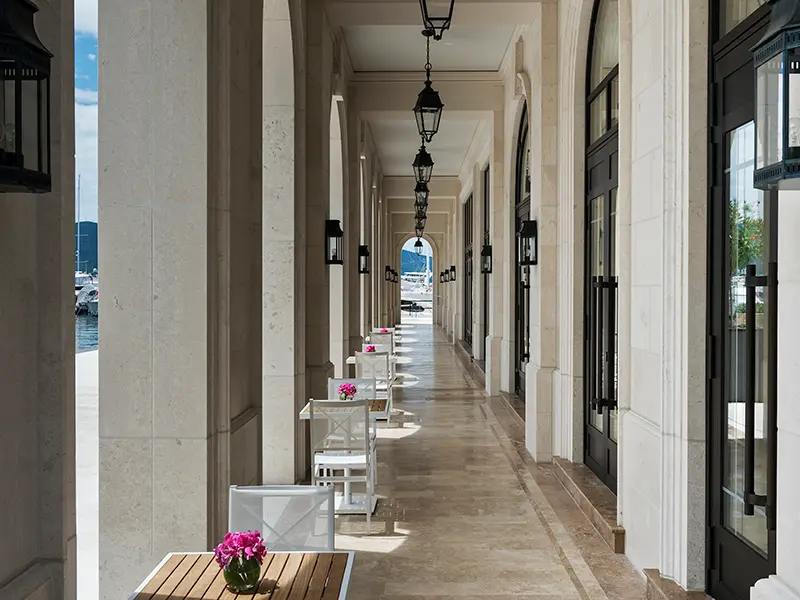 Arkadni hodnik u Regent Porto Montenegro sa kamenim stubovima, crnim lampionima, belim stolovima i stolicama i pogledom na marinu