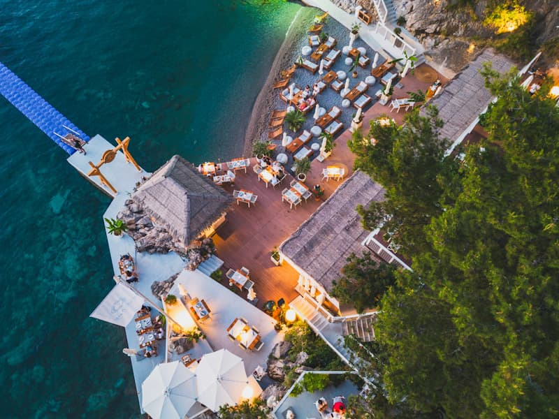 Aerial večernji pogled na platformu Escape Plaža i Restoran sa thatched krovom bara, stolovima pod suncobranima, molom i tirkiznim morem