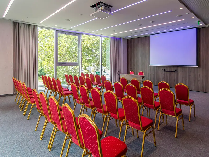 Konferencijska sala u Hotel Sheraton Novi Sad sa teatarskim rasporedom, crvenim stolicama sa zlatnim okvirom, projektorom, platnom i panoramskim prozorima