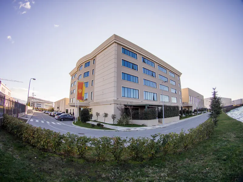 Panoramski pogled na fasadu Hotel voco Podgorica sa bež fasadom, zastavom Crne Gore, zelenim živicama, parkingom i plavim nebom