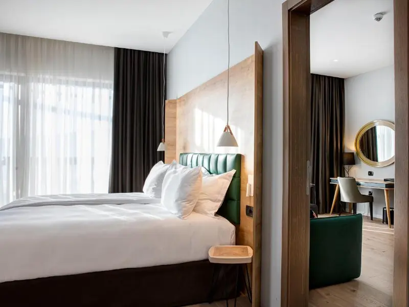 Apartman u Tarčin Forest Resort & Spa Sarajevo hotelu sa bračnim krevetom, zelenim uzglavljem, visećim lampama i pogledom na dnevnu sobu kroz otvor