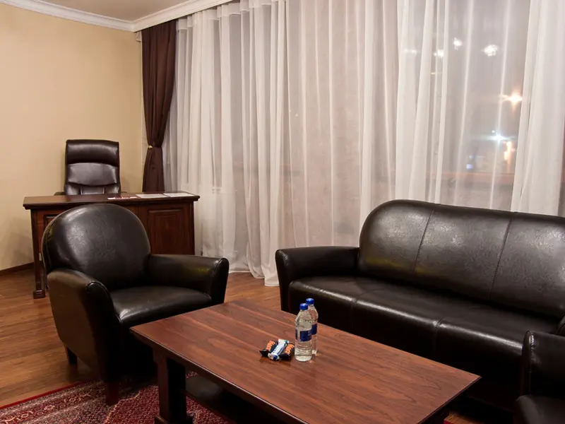 Dnevna soba apartmana u Hill Hotel sa crnim kožnim foteljama i kaučem, drvenim radnim stolom, persijskim tepihom i braon zavesama