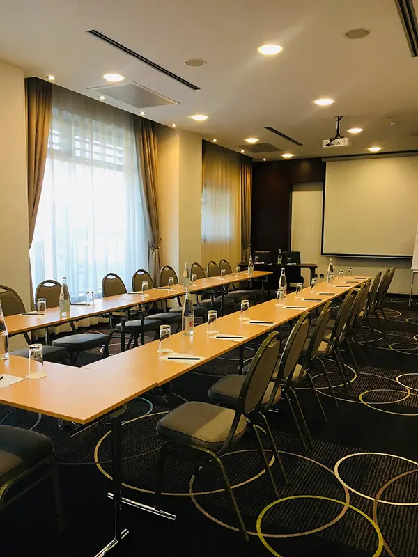 Konferencijska sala u IN Hotel sa stolovima u U-rasporedu, projektorskim platnom, tamnim tepihom sa geometrijskim motivima i zlatnim zavesama
