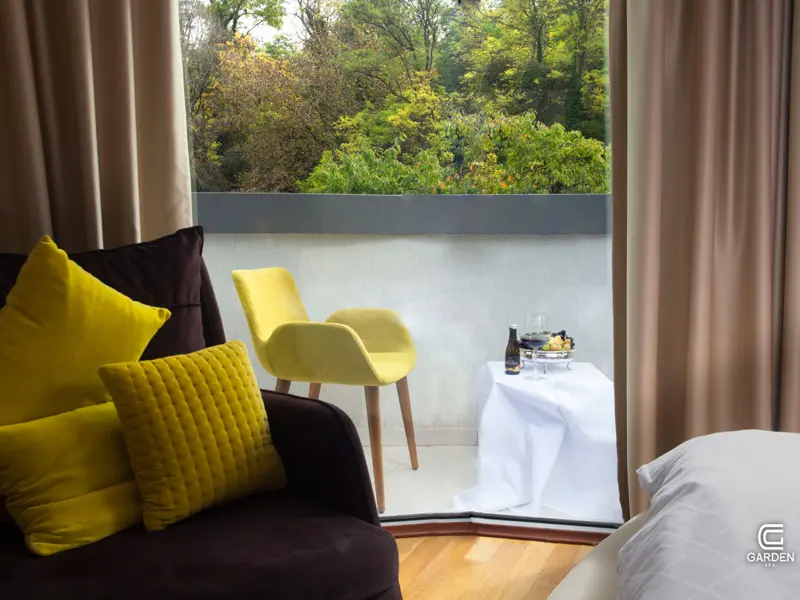 Detalj sobe u Garden Spa Boutique Hotel sa tamnom foteljom, žutim jastucima, žutom stolicom na balkonu, šampanjcem i pogledom na zelenilo