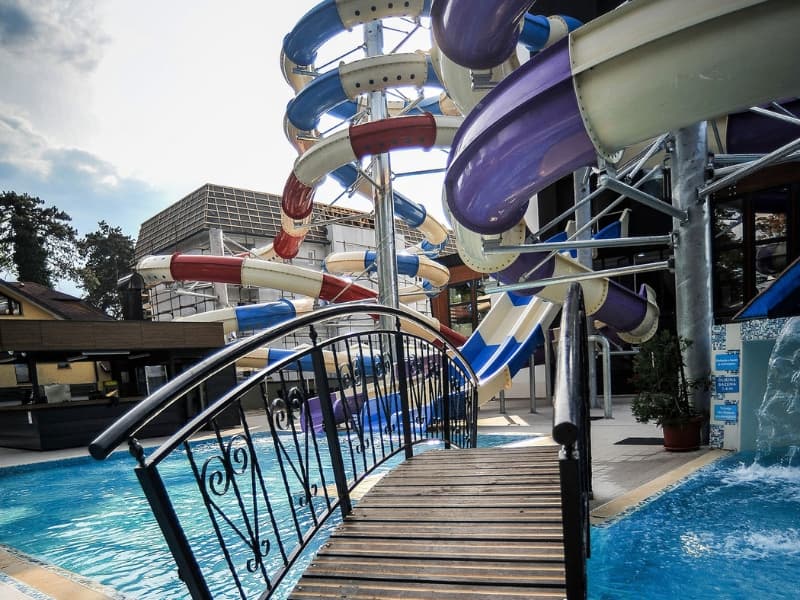 Akva park u Hotel Vrnjačke Terme sa šarenim tobogan stazama, drvenim mostom preko bazena, metalnom ogradom i pogledom na hotel