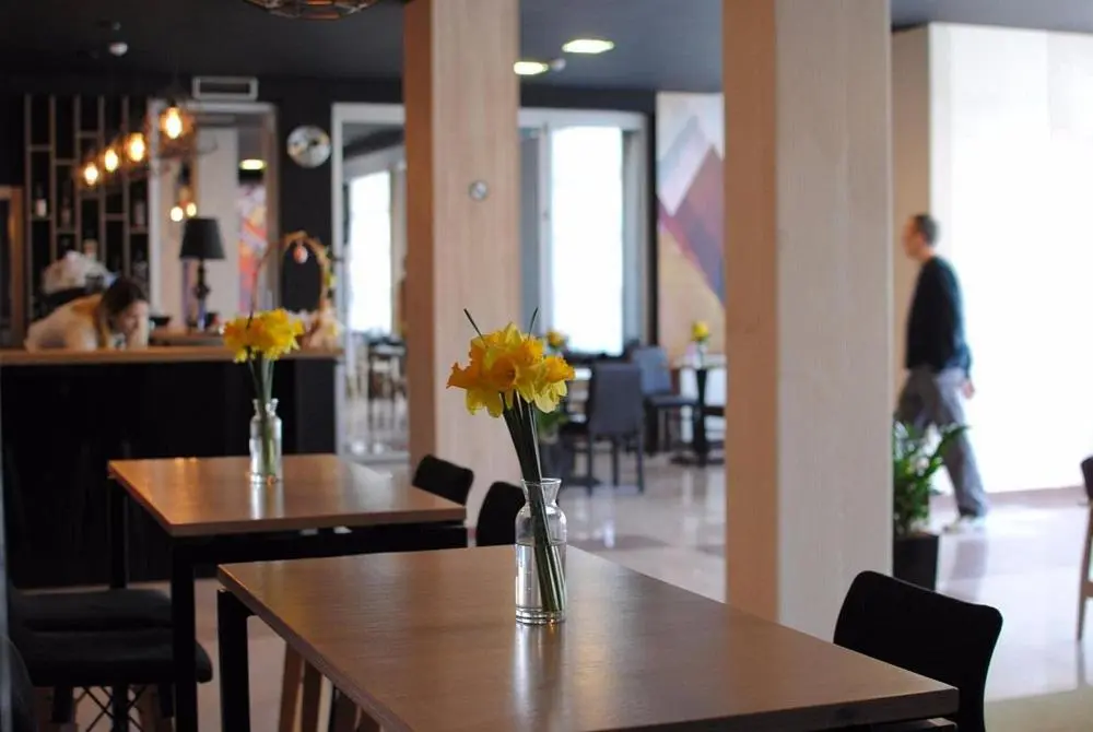 Kafić u Hotel Porta sa drvenim stolovima, žutim narcisima u vazama, modernim visećim lampama i šarenim umetničkim zidom