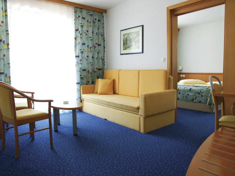 Dnevna soba apartmana u Ramada Resort Kranjska Gora hotelu sa žutim kaučem, stolicama, šarenim zavesama i pogledom na spavaću sobu