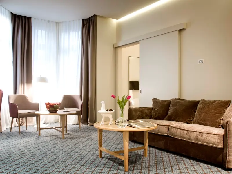 Dnevni boravak apartmana u Jump Inn Hotel Belgrade sa braon plišanim kaučem, foteljom, okruglim stolićem, plavim tepihom i tulipanima