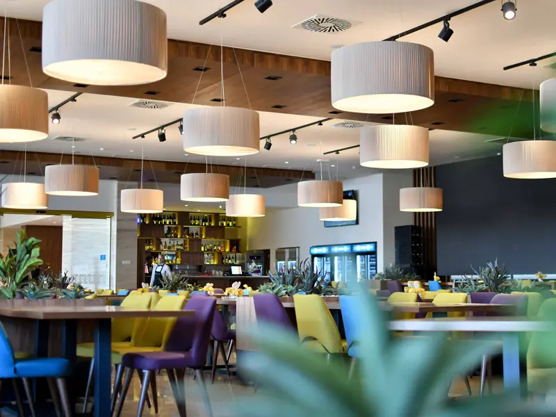 Bar i restoran u Hotel Nais sa cilindričnim visećim lampama, barom sa osvetljenim policama, šarenim stolicama i biljkama kao dekorativnim pregradama