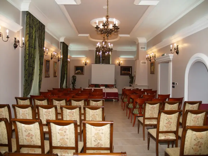 Konferencijska sala u Lovačkom Dvorcu Kaštel Ečka sa drvenim stolicama u redovima, projektorskim platnom, bronzanim lusterom i zelenim zavesama