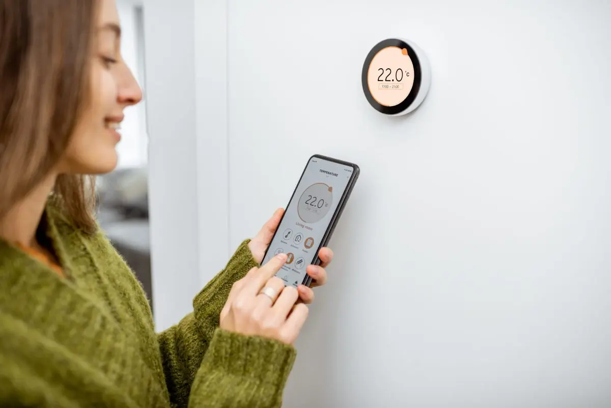 programmable thermostat installation Woman dressed in green sweater regulating heating temperature with a modern wireless thermostat and smart phone at home. Synchronization of thermostat with mobile devices concept