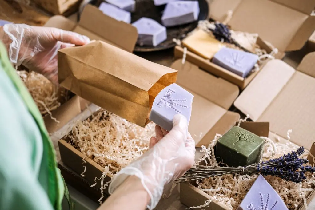 indie craft parade greenville Person packs lavender soap into a kraft paper bag surrounded by shredded filler and handmade bars. Handmade product shipping, indie beauty dispatch, craft brand logistics, eco-packaging workflow.