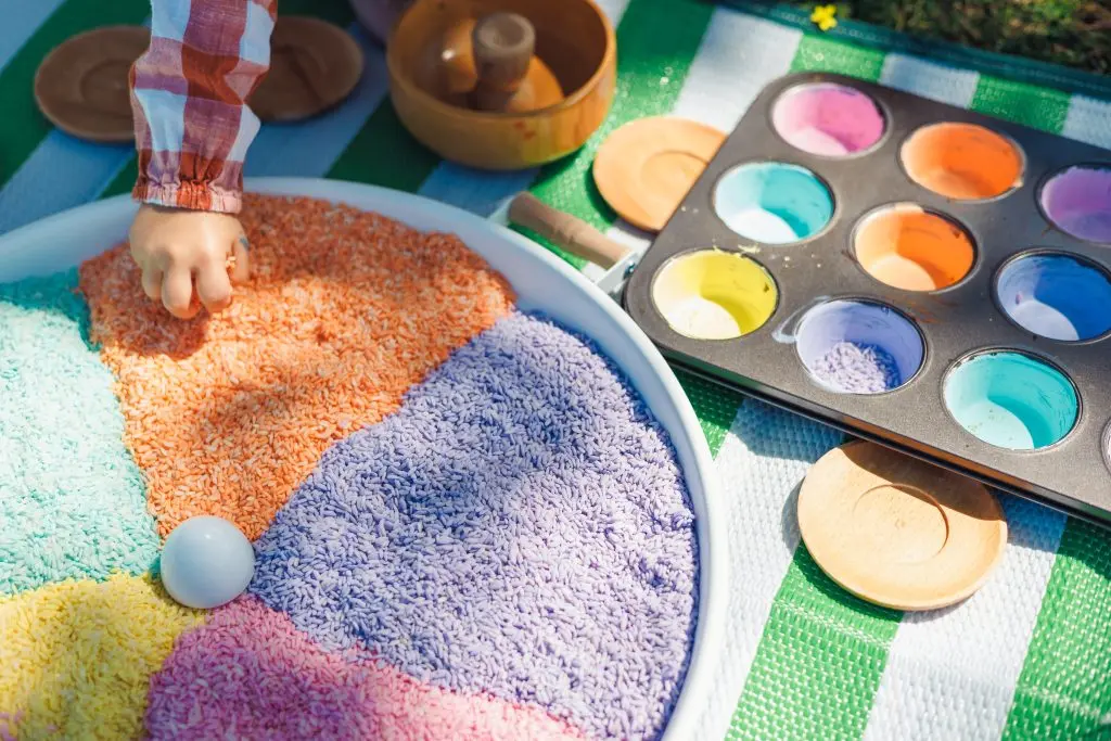 indie craft parade greenville Children engage in creative sensory play by pouring colored rice into sections of a large tray while surrounded by colorful paint. The setting is a sunny park with green grass.