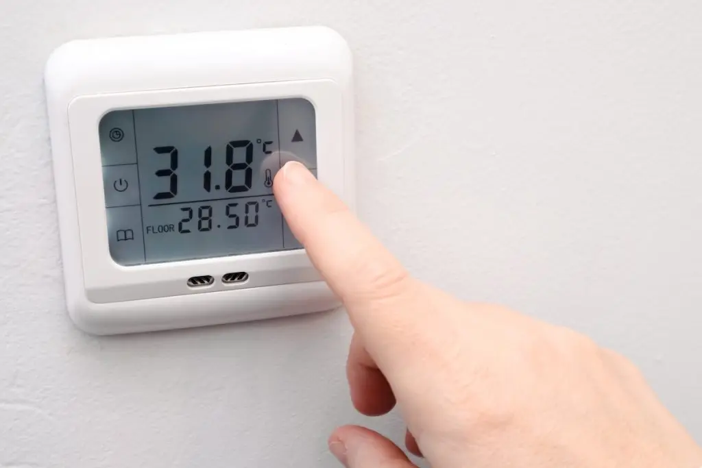 Close-up of a finger pressing a button on a white electronic thermostat. The screen displays a temperature of 31.8°C, representing smart home climate control and energy efficiency