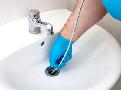 Hand in blue glove using a drain snake to unclog a sink, highlighting professional drain cleaning services for plumbing maintenance.