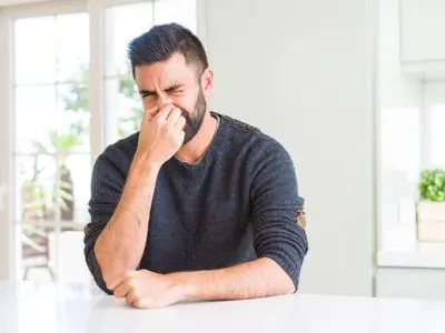 Man sitting at a table, grimacing and holding his nose, indicating a bad smell, relevant to HVAC duct issues and indoor air quality concerns.