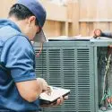Technician servicing an air conditioning unit with a diagnostic tablet, tools, and open panel at Carmine's Plumbing.