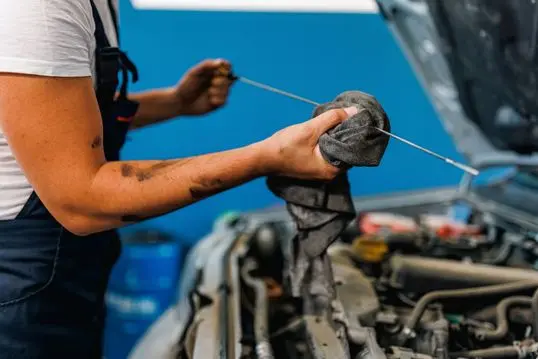 A close-up picture of a young Caucasian male mechanic standing next to a car with a popped hood and wiping a car part from motor oil, using a rug.