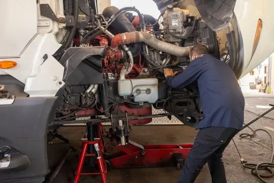 A male mechanic in blue work attire focuses on repairing a truck engine in a professional garage. Tools and equipment surround the vehicle on a hydraulic lift.