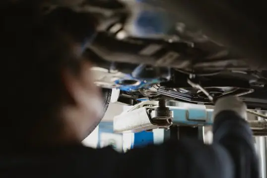A mechanic is focused on repairing a vehicle from underneath, with various tools visible around. The setting appears to be a professional repair shop.