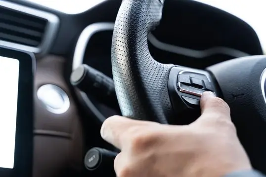 Close-up of driver hand pressing multifunction steering wheel button inside modern vehicle, highlighting automotive technology, comfort, and safe driving concept for transportation imagery use.