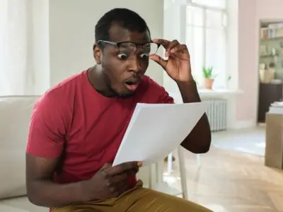 Surprised man reading document, expressing shock at increased energy bills, in cozy home setting.