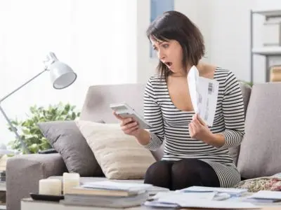 Woman sitting on a couch, shocked while reviewing a high energy bill and using a calculator, illustrating concerns about rising heating costs and the need for heater repair.