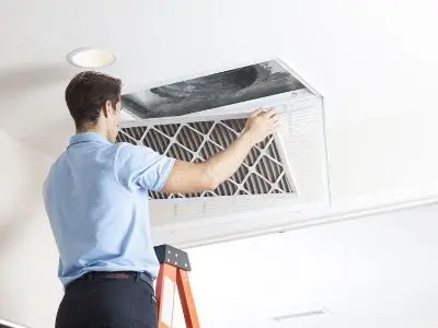 Man replacing an air filter in a ceiling-mounted air conditioning unit, illustrating essential AC maintenance tips for homeowners.