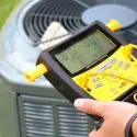 Technician using a digital HVAC gauge to assess air conditioning system performance, emphasizing the importance of maintenance before summer heat.