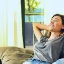 Woman relaxing on a couch, enjoying cool air in a well-lit living room, emphasizing comfort and relaxation in a home environment.