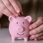 Person inserting a coin into a smiling pink piggy bank, symbolizing savings and energy cost reduction related to HVAC services.