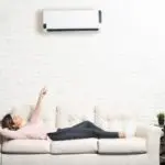 Woman relaxing on a couch pointing at a ductless mini-split air conditioning unit on the wall, illustrating comfortable indoor air quality and HVAC solutions.