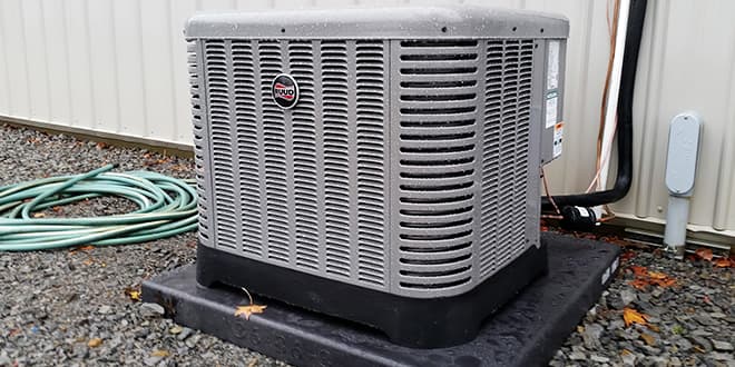 HVAC unit on a black base outside a metal building with green garden hose nearby.