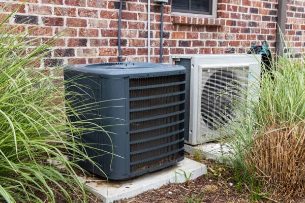 Outdoor air conditioning units next to a brick wall, surrounded by grass plants.