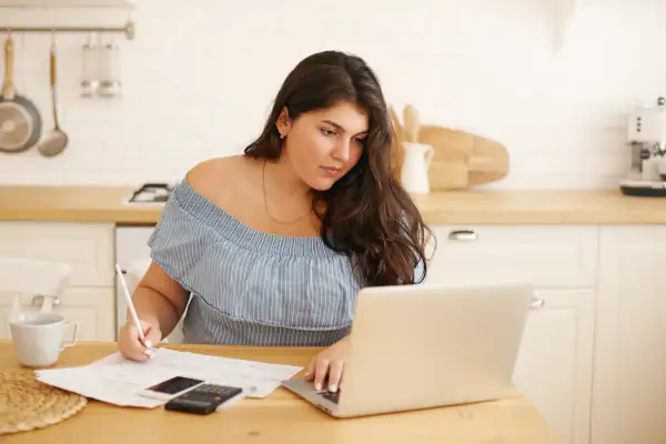 The Advantages of Energy-Efficient AC Systems: Why They're a Smart Investment - Young woman staring at an open laptop while taking notes next to a handheld calculator.