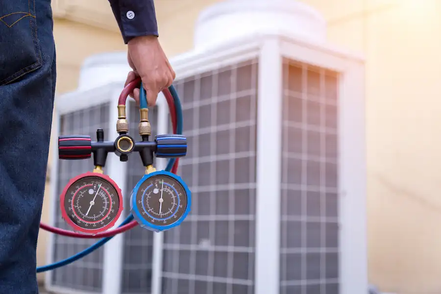Technician holding HVAC manifold gauges near an outdoor air conditioning unit.