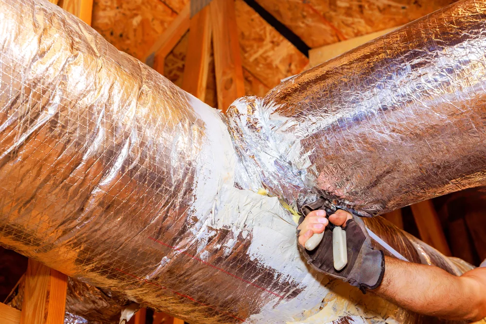 A person wearing gloves seals ductwork with foil tape in an attic space.