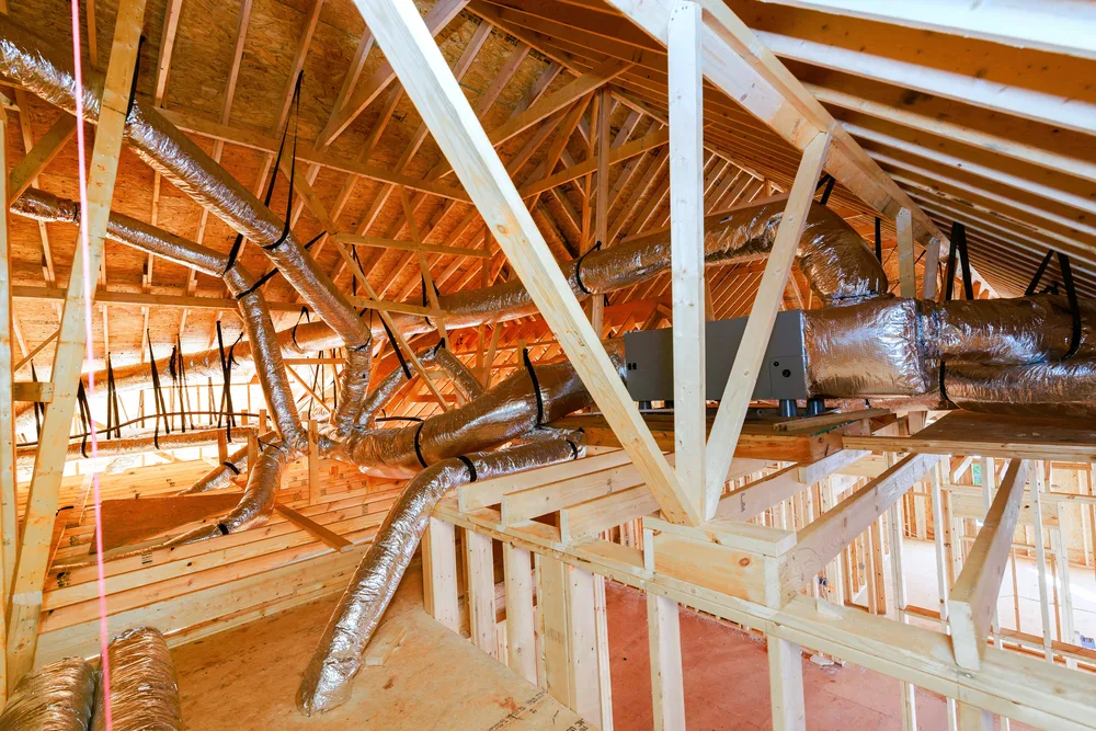 Exposed HVAC ductwork with silver insulation in an unfinished attic space with wooden rafters.