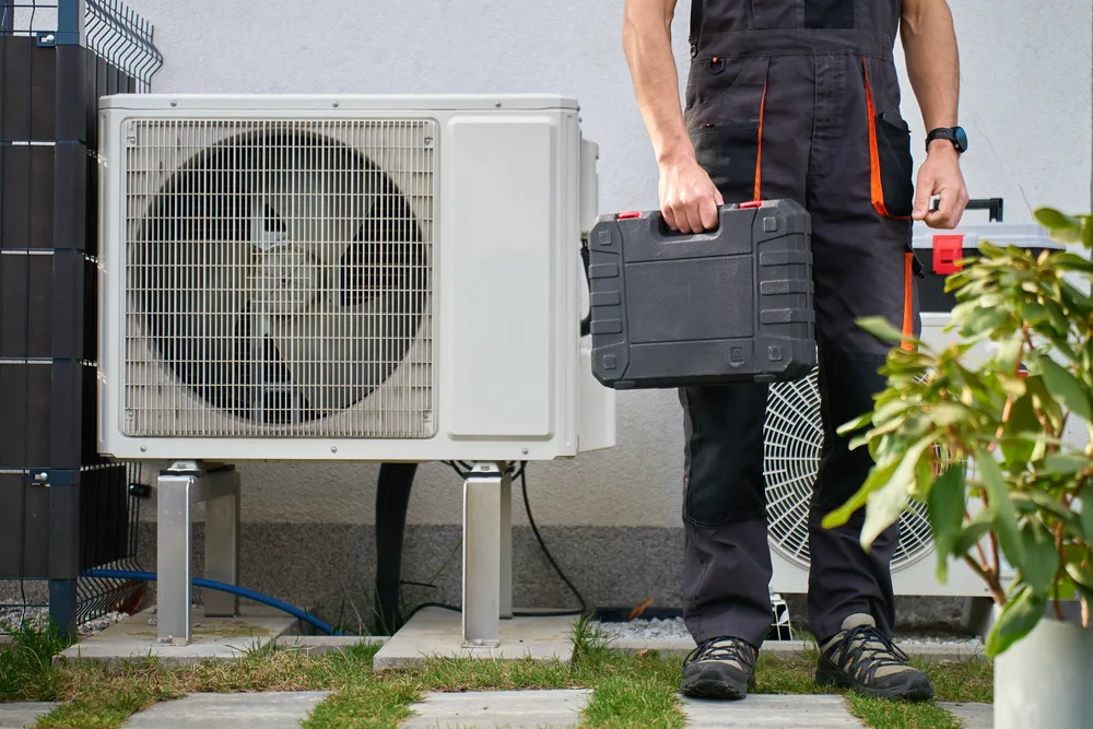 Technician in uniform holding a toolbox next to outdoor HVAC units.