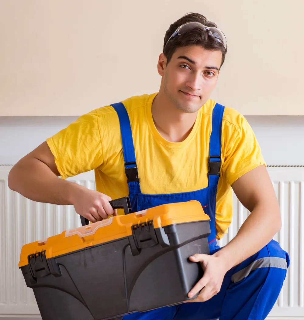 Technician in blue overalls and a yellow shirt holds a large toolbox.