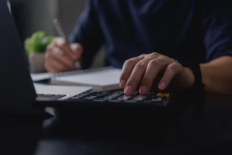 Hand using a calculator with a blurred notebook and pen in the background.