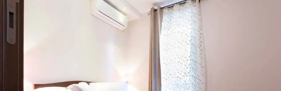 Bedroom featuring an air conditioning unit above a bed, with natural light filtering through a decorative curtain, illustrating effective cooling solutions for individual rooms.