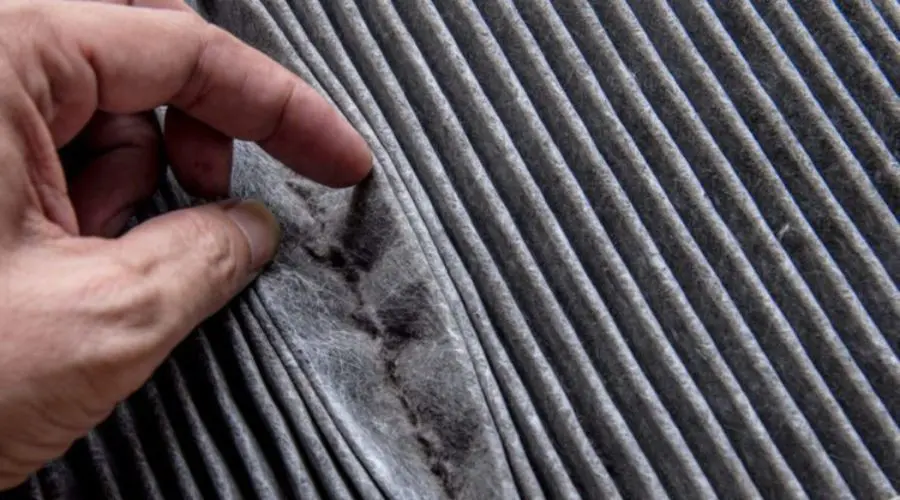 A dirty, pleated air conditioning filter being held and displayed by a hand. The filter shows accumulated dust and debris.