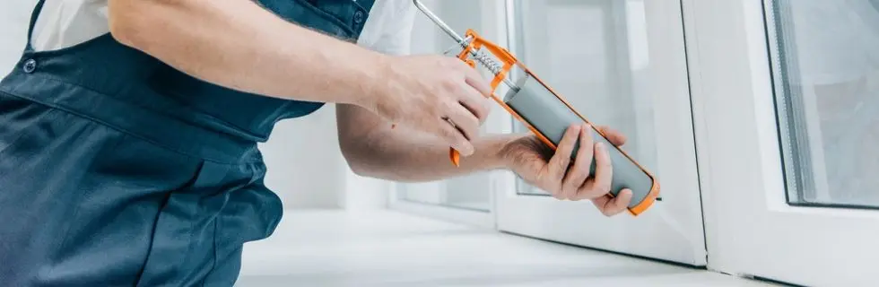 Person applying caulk to seal air leaks around a window, demonstrating a DIY method for improving home energy efficiency and comfort.