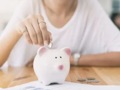 Person inserting a coin into a piggy bank on a wooden table, symbolizing savings and financial management.