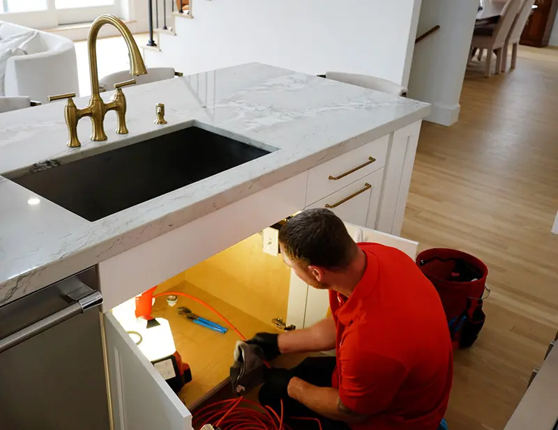 Plumber in red shirt working under kitchen sink, showcasing plumbing services for HVAC and plumbing needs in Taunton, MA.