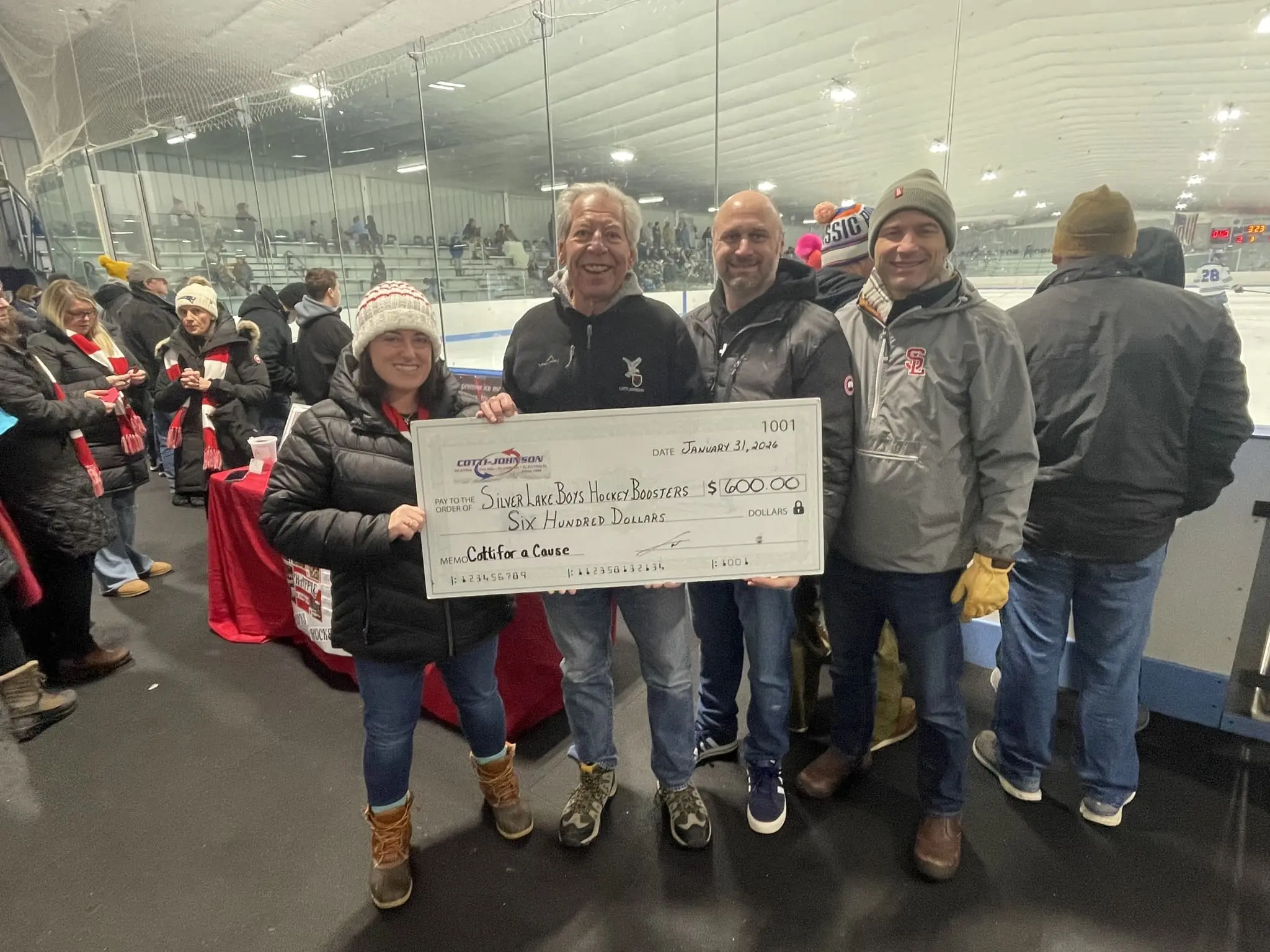 Cotti-Johnson HVAC representatives presenting a $600 check to Silver Lake Hockey Boosters during a community event, with supporters in the background at an ice rink.
