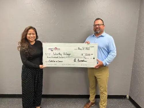 Mayra Perez and Adam V. Boccalini holding a large check for $500 donation to Worthy Village, symbolizing Cotti-Johnson's support for the nonprofit assisting Guatemala.