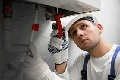 HVAC technician inspecting water heater components with a red tool, wearing gloves and a hard hat, in a home setting.
